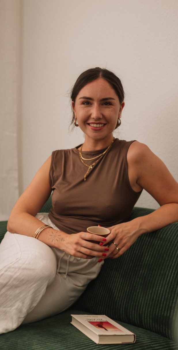 Das Bild zeigt Giorgia Nebuloni, eine Heilpraktikerin für Psychotherapie aus Köln, vor einem Holzzaun, der von üppigem, grünem Laub und Pflanzen umgeben ist. Sie trägt ein schwarzes Kleid und lächelt sanft. Giorgia hat lange, dunkle Haare und trägt goldene Ohrringe. Die natürliche und beruhigende Umgebung reflektiert ihre einfühlsame und harmonische Herangehensweise in der Psychotherapie.