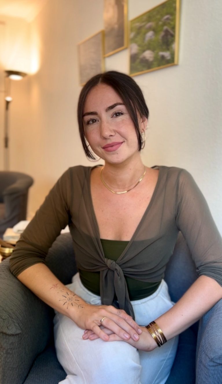 Das Bild zeigt Giorgia Nebuloni, eine Heilpraktikerin für Psychotherapie aus Köln, in einem grünen Garten sitzend. Sie trägt ein schwarzes Kleid und lächelt freundlich. Giorgia sitzt auf einer Holzbank, umgeben von verschiedenen Pflanzen und Blumen. Die entspannte und natürliche Umgebung strahlt Ruhe und Harmonie aus, was ihre therapeutische Philosophie widerspiegelt.